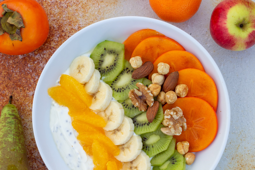 Frugtbowl med appelsin, banan, kiwi, sharonfrugt og nødder.